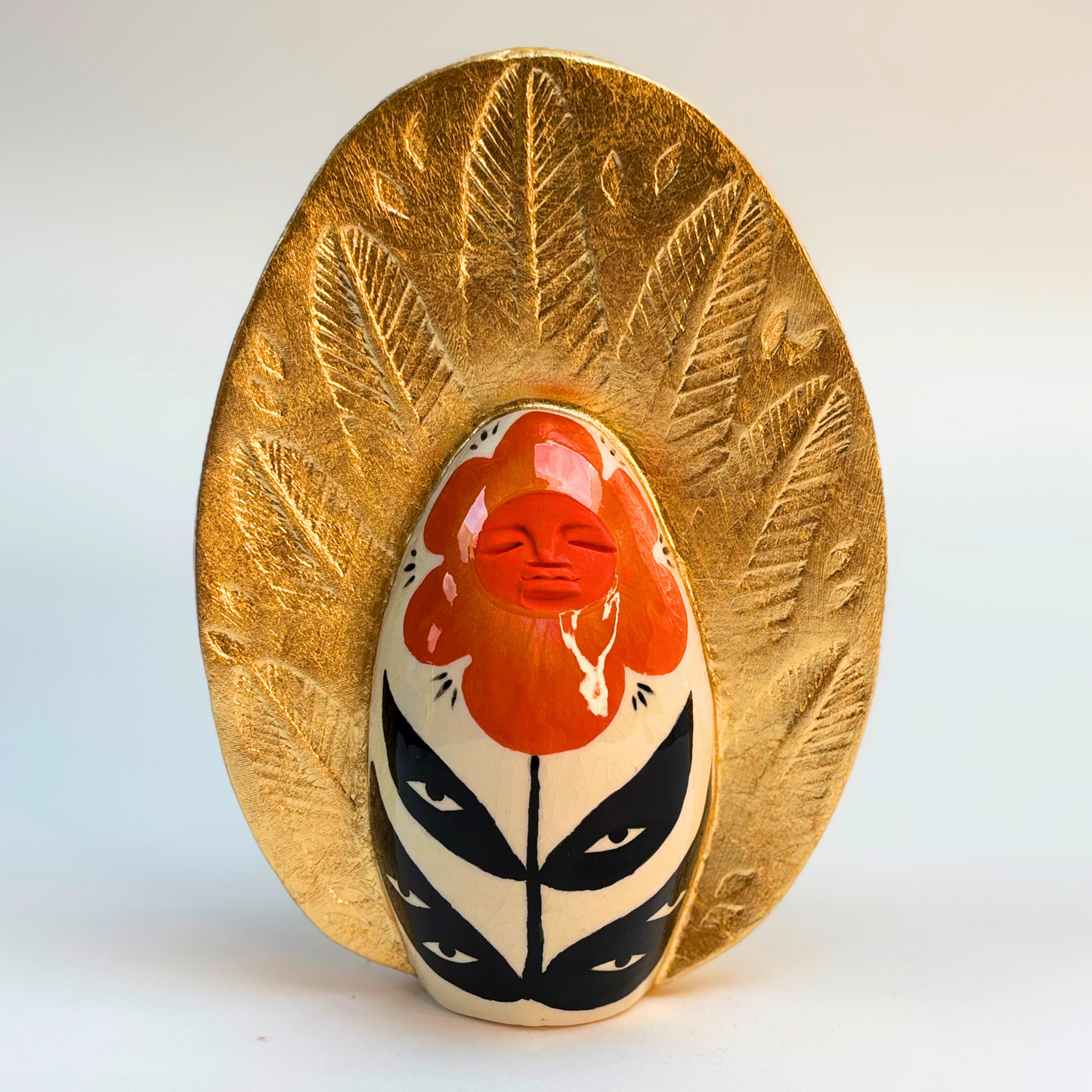 front view of ceramic goddess geode with orange petal peaceful face on a cream ground with black leaves and eyes on them with a golden crown with embossed leaves in the gold leaf crown