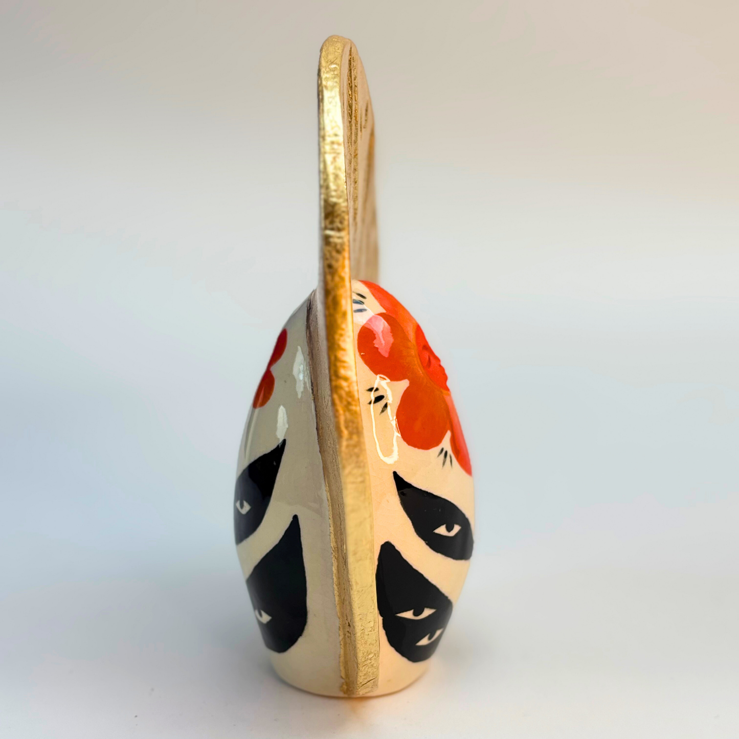 side view of ceramic goddess geode with orange petal peaceful face on a cream ground with black leaves and eyes on them with a golden crown with embossed leaves in the gold leaf crown