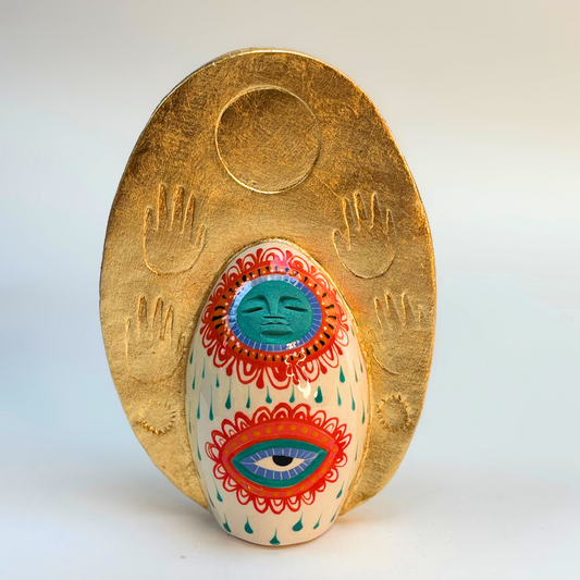 front view of gold crown of goddess surrounds the  cream ground body of goddess with turquoise face, circled by blue, red and orange patterns with an eye beneath and turquoise tear drops on the body