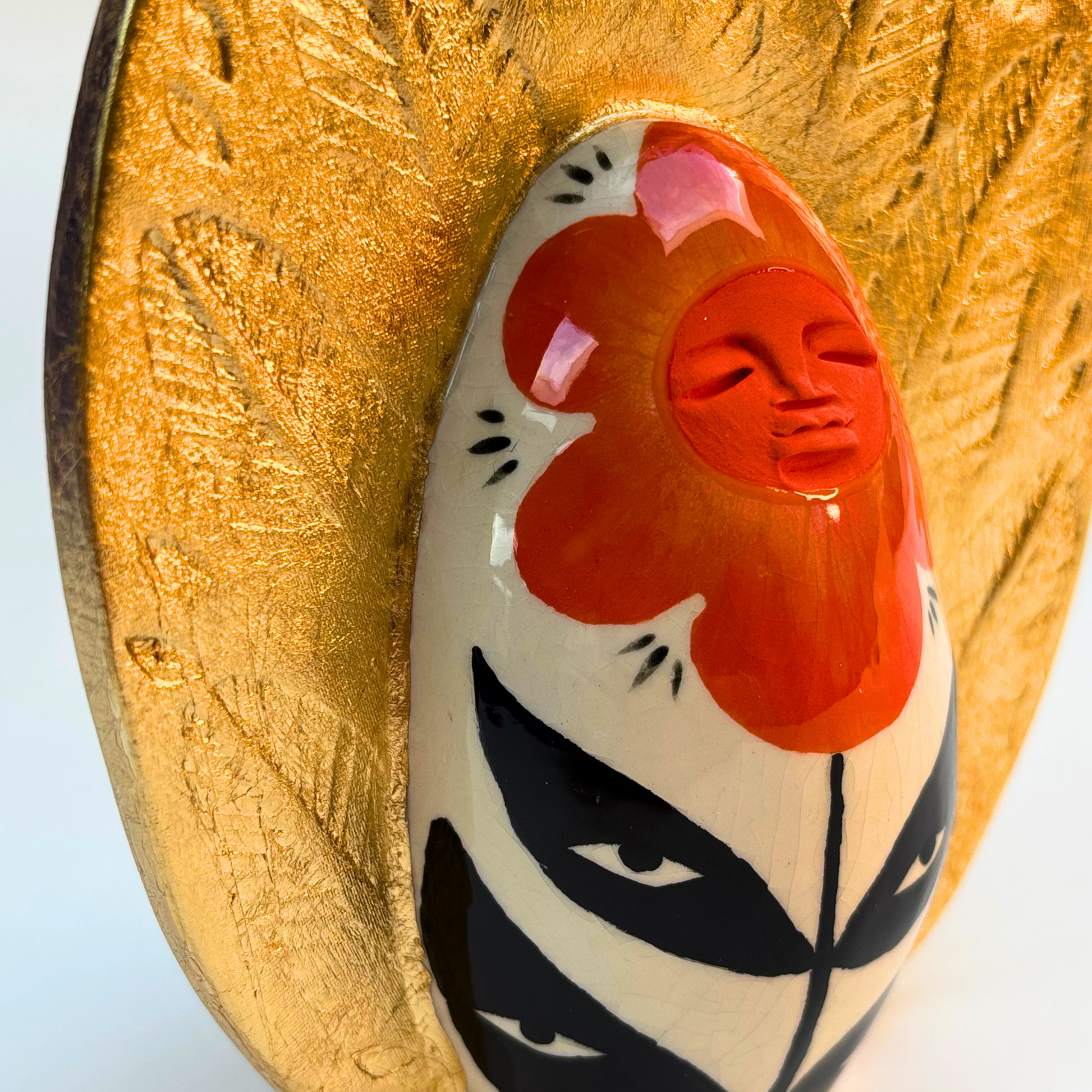 close up of a peaceful flower face in orange of a ceramic goddess geode on a cream ground with black leaves and eyes on them with a golden crown with embossed leaves in the gold leaf crown